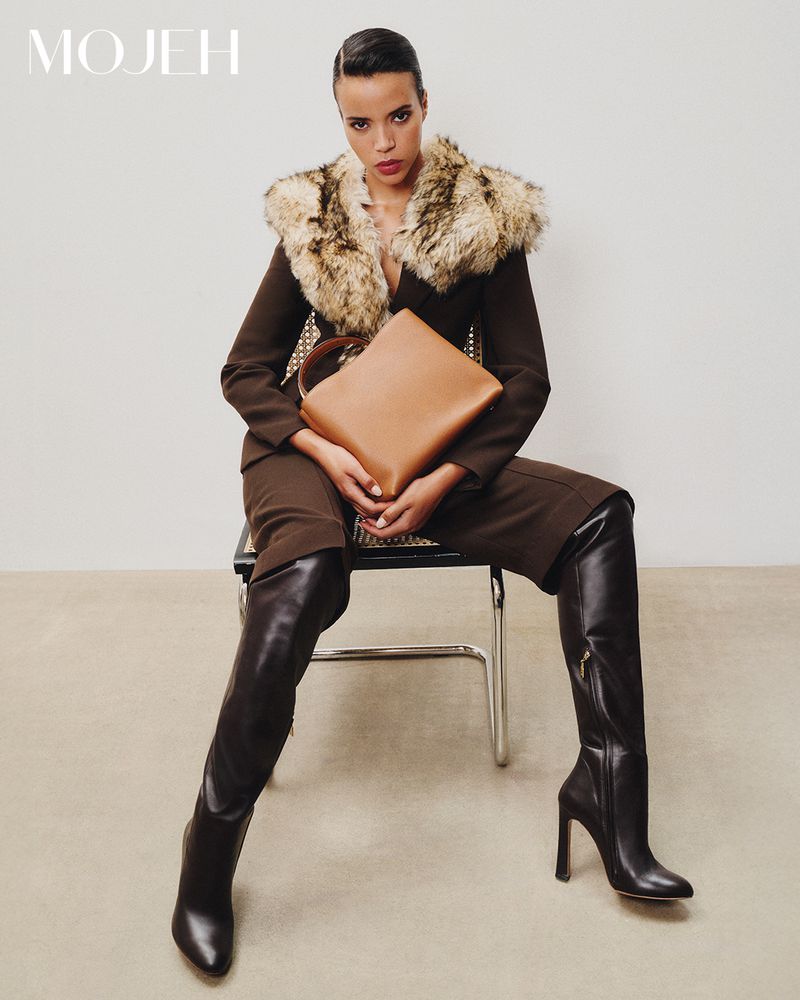 Model Calista Choley seated in a brown tailored outfit with fur collar and black knee-high boots holding a tan leather bag.