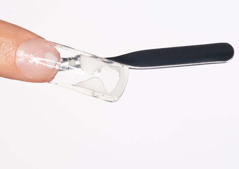 Close-up of a fingernail with a clear artificial nail form attached and a metallic nail tool on a white background. Beauty shot.