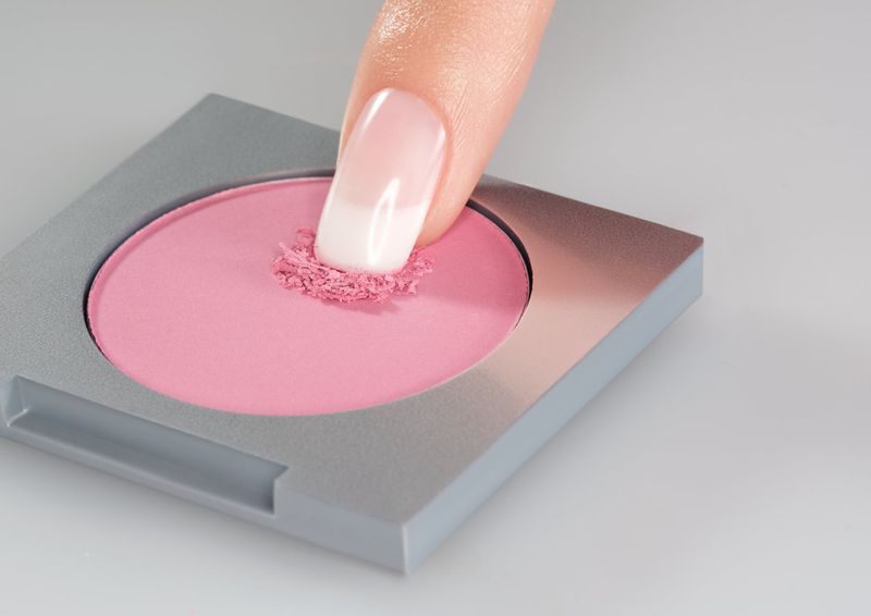 Close-up of a finger pressing into pink blush powder in a gray compact, focusing on the makeup and application for beauty concepts.