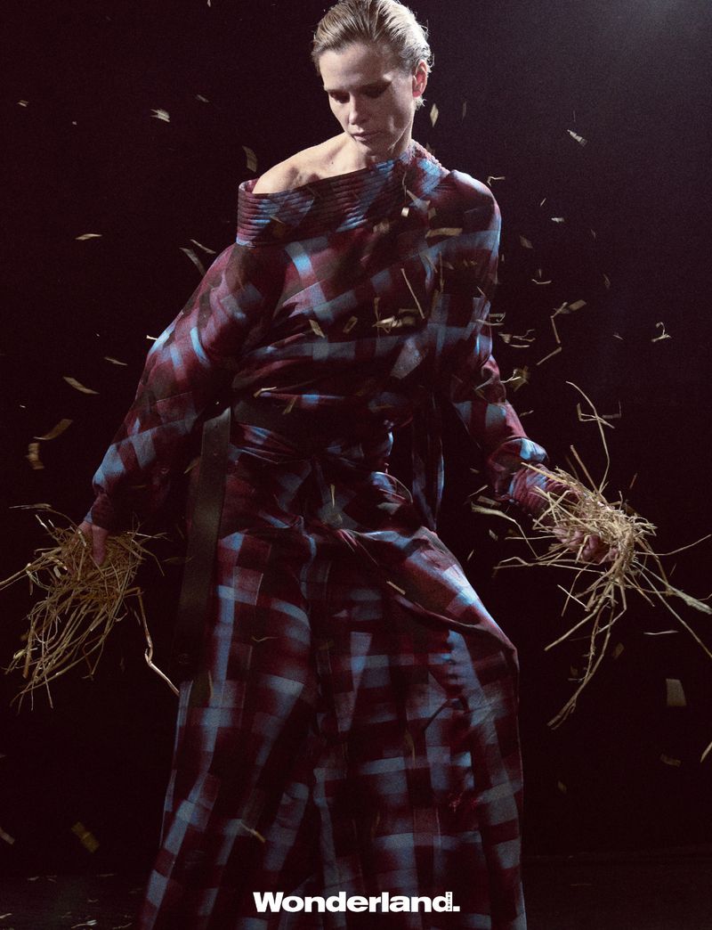 Fashion shot of a blonde woman in a checkered dress holding straw, with dark background and falling straw-like objects for an editorial look.