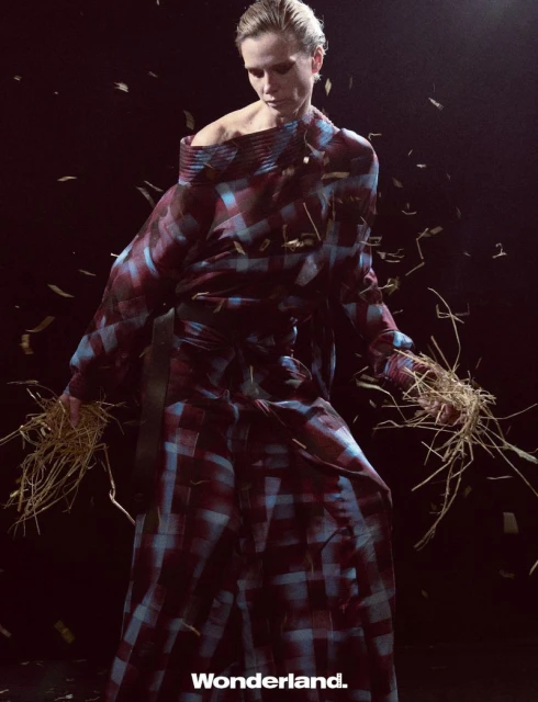 Fashion shot of a blonde woman in a checkered dress holding straw, with dark background and falling straw-like objects for an editorial look.