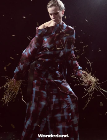Fashion shot of a blonde woman in a checkered dress holding straw, with dark background and falling straw-like objects for an editorial look.