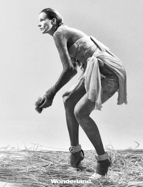 Fashion shot of an adult woman in dress, high heels, and a crouched pose, standing on hay in a photo studio with a light grey backdrop.