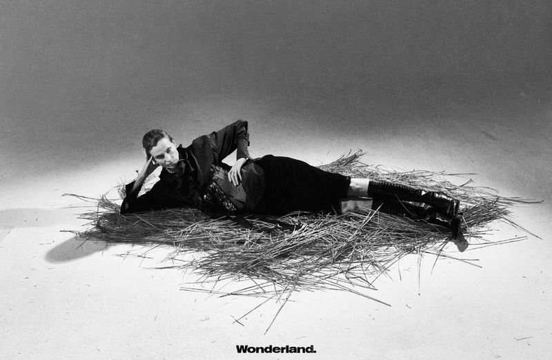 Monochrome studio shot of a person in dark clothing reclining on a bed of straw, striking a pose, with a light grey backdrop.