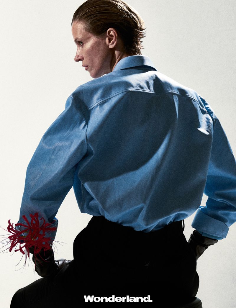 Woman in a light blue button-down shirt and black pants, seated, with red spider lilies in her hand, in a fashion editorial.
