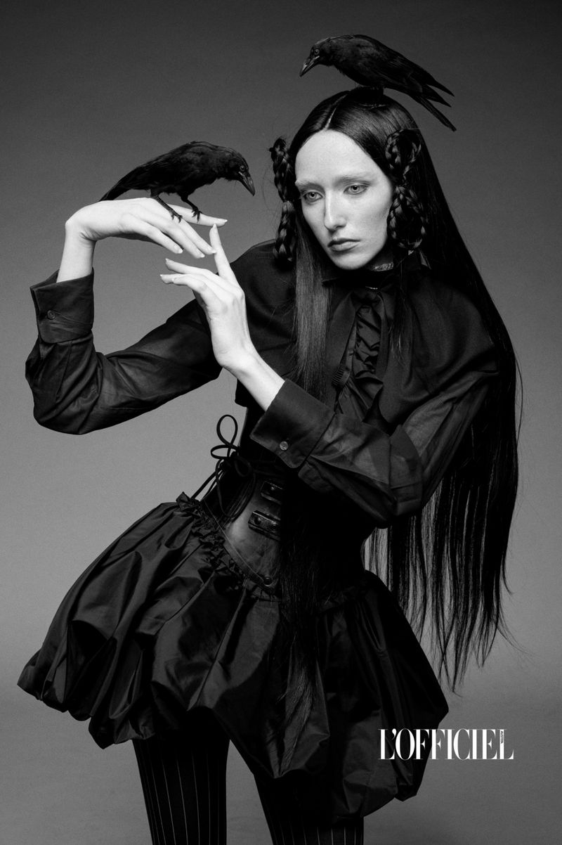 Striking black-and-white fashion shot featuring a model with long black hair, dark clothing, and two ravens, set against a plain backdrop.