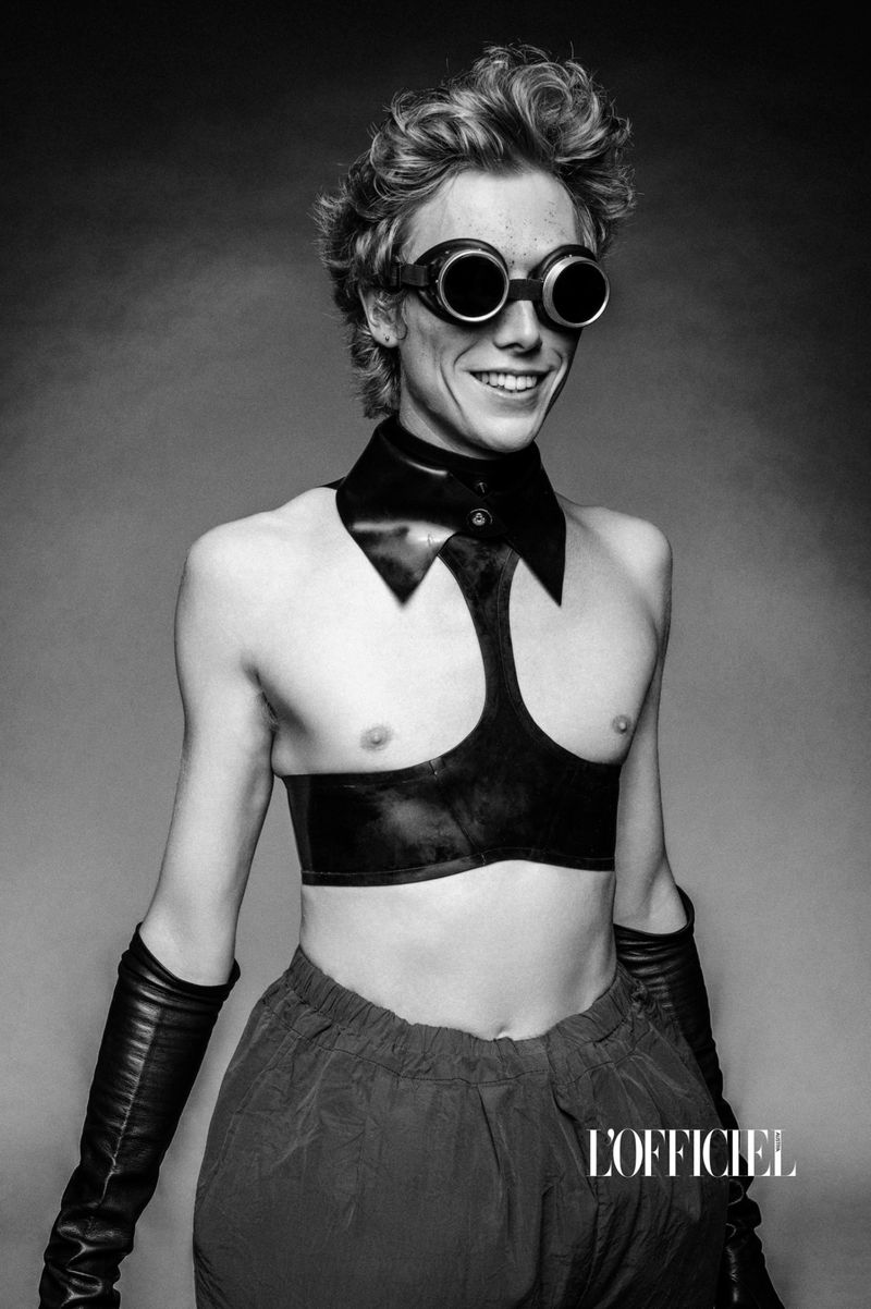 A person with goggles and an open top smiles in a studio shot. They wear long black gloves and reddish pants against a grey backdrop.