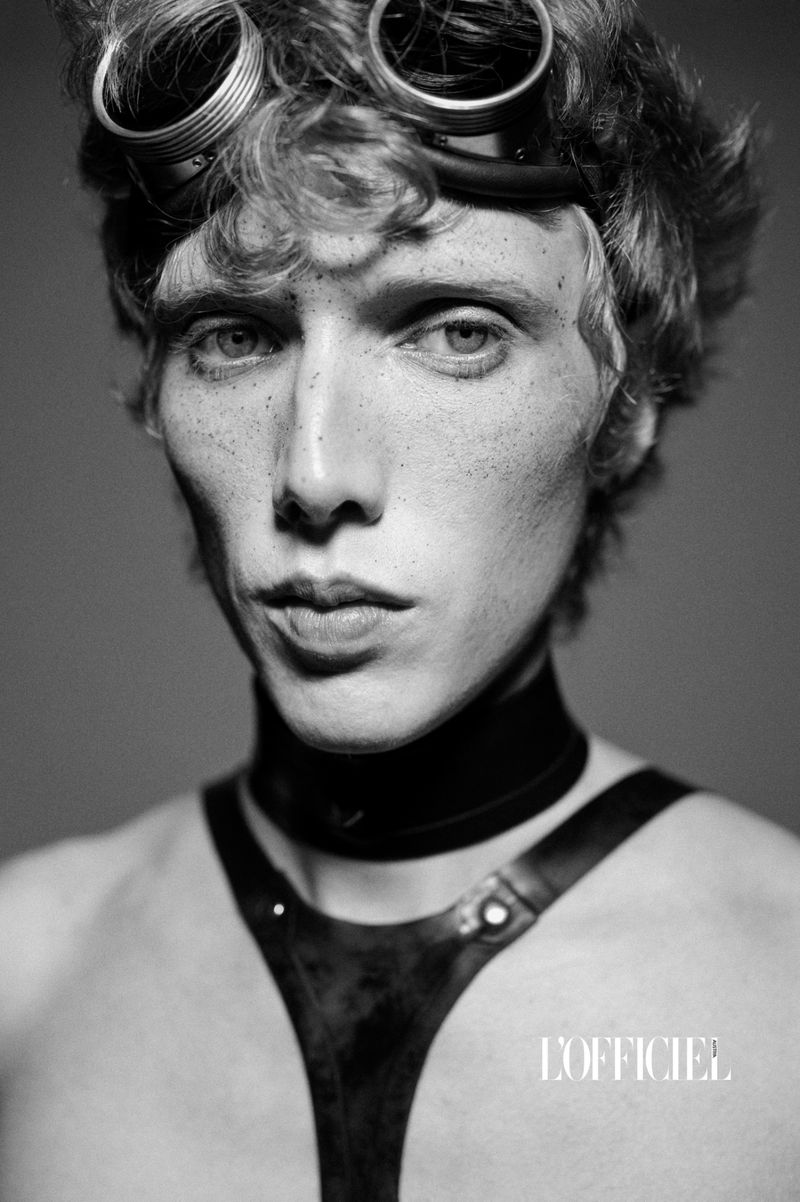 Black and white studio portrait of a young adult male with goggles on his head, wearing a leather harness and choker, looking at the camera.