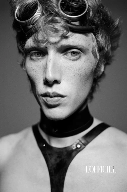 Black and white studio portrait of a young adult male with goggles on his head, wearing a leather harness and choker, looking at the camera.