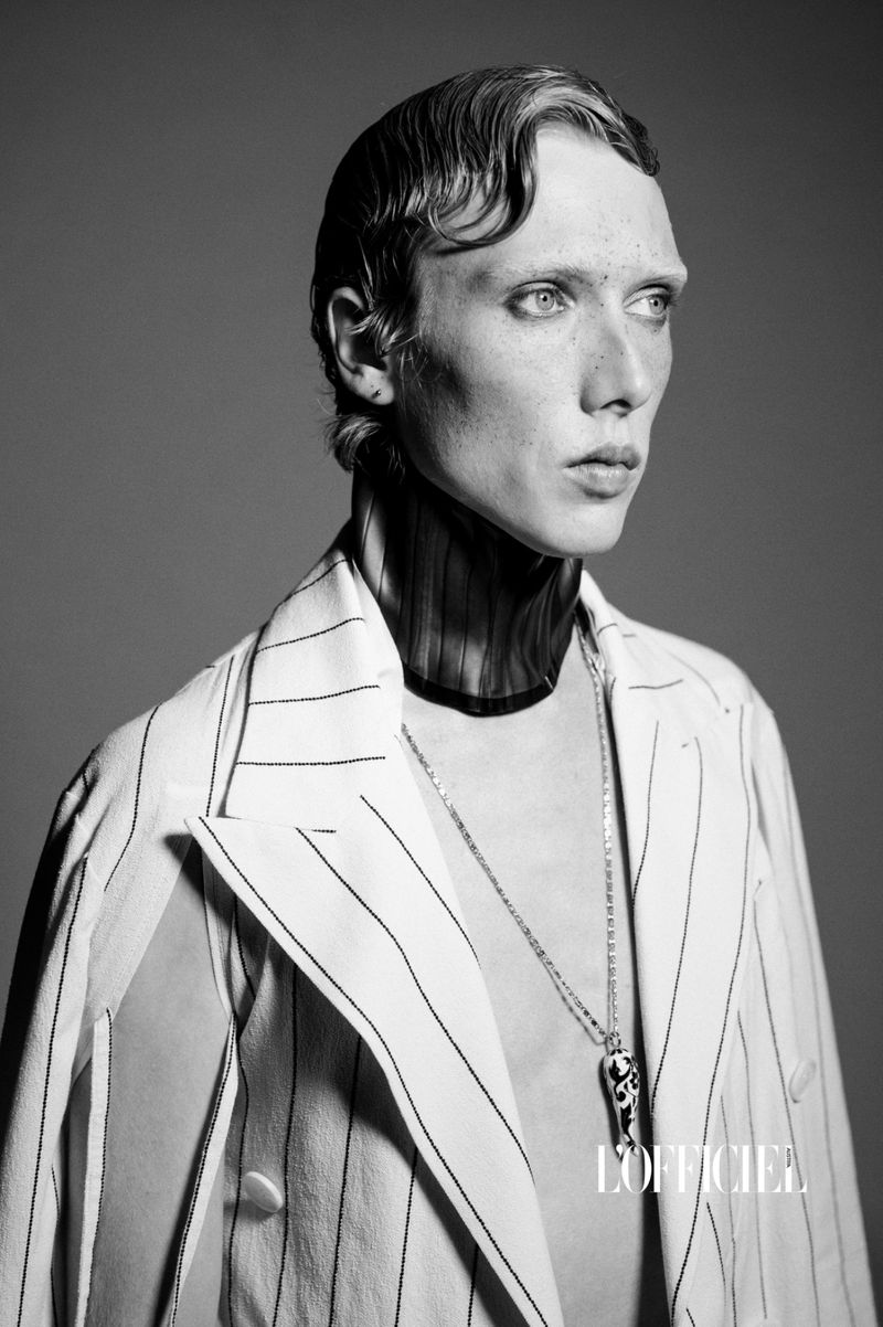 Black and white portrait of a slender male model in a pinstripe suit with pendant against a grey background in a studio setting.