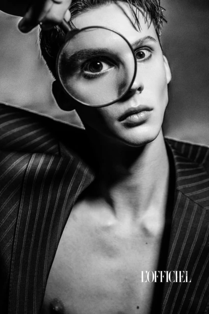Monochrome portrait of a young man with fair skin looking through a magnifying glass, wearing a pinstripe jacket in a studio setting.