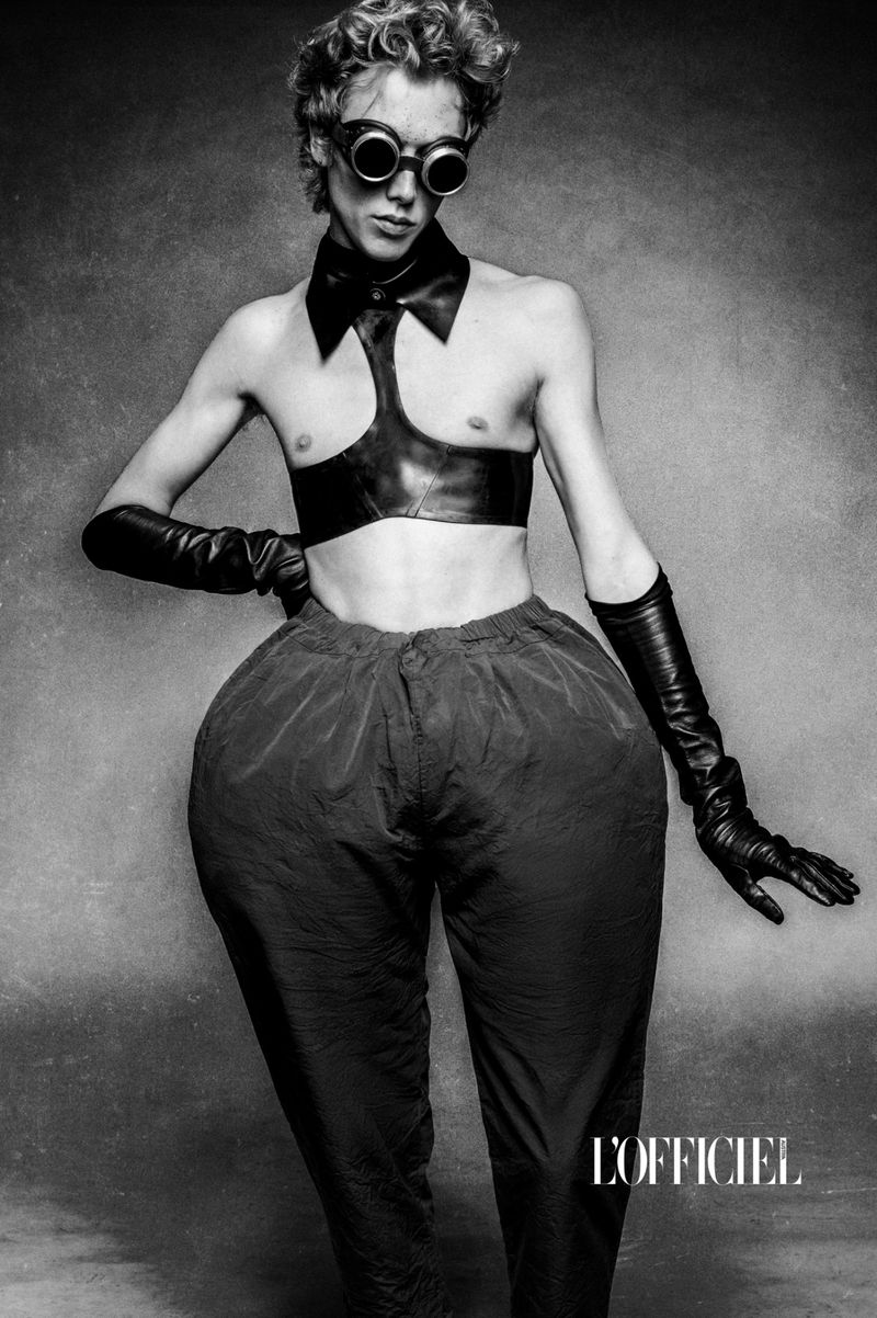 Monochrome portrait of a slender person in avant-garde fashion, leather top, oversized pants, gloves, and goggles against a textured gray backdrop.