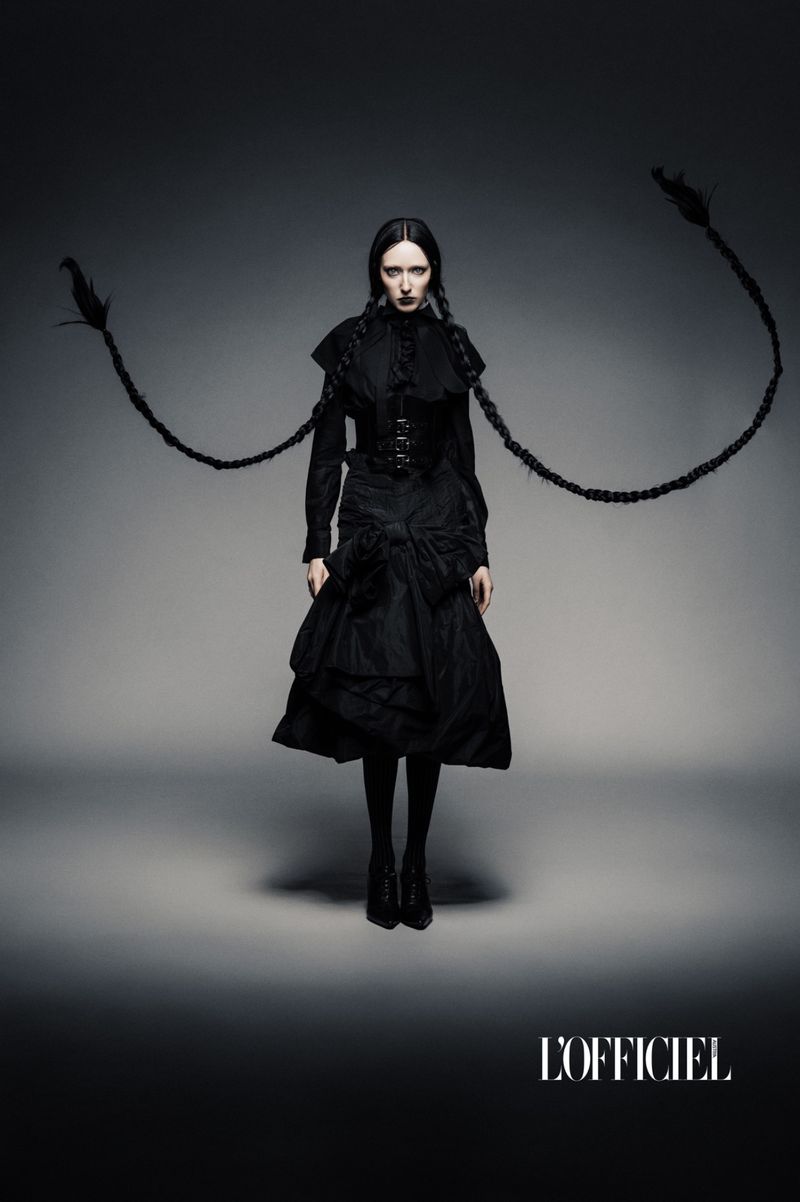 Fashion editorial portrait of a woman with long braids in a black gothic-inspired dress against a dark grey gradient backdrop.