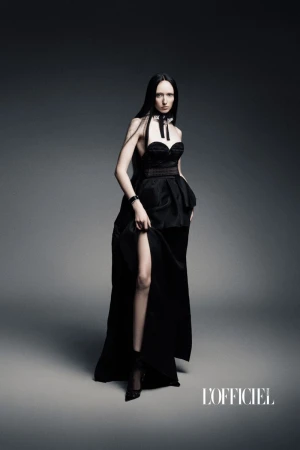 A model poses in a black gown with a high slit against a gray background in a studio. The image is a fashion editorial.