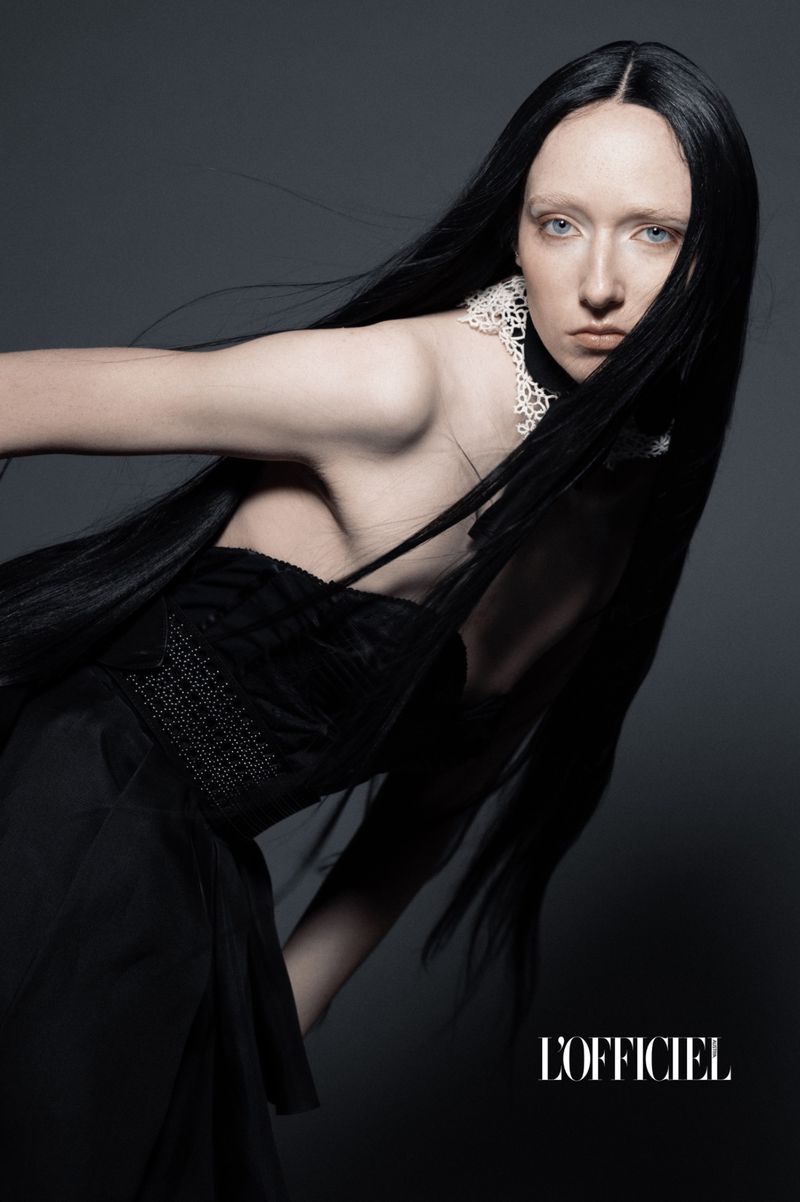 Portrait of a woman with long black hair wearing a black dress and lace collar posing in a studio with dark grey background and 'L'Officiel' logo.