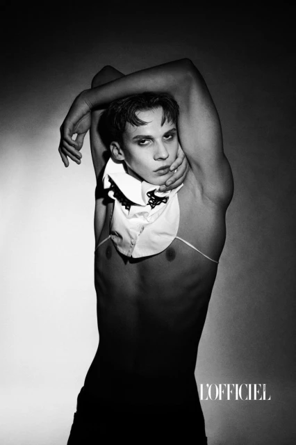 Black and white portrait of a shirtless male model wearing a decorative collar in a studio setting with soft, diffused lighting.