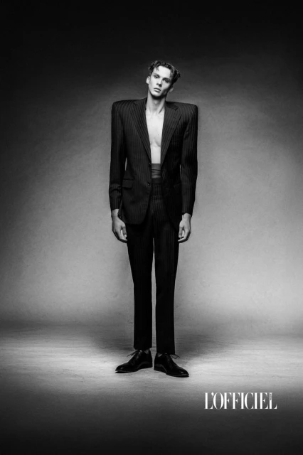Monochrome fashion shot of a slender man in a pinstripe suit with an open jacket, standing in a photo studio, against a neutral gray backdrop.