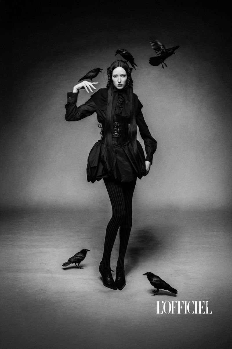 A fashion editorial shot in black and white, featuring a woman in dark clothing surrounded by ravens in a neutral studio setting.
