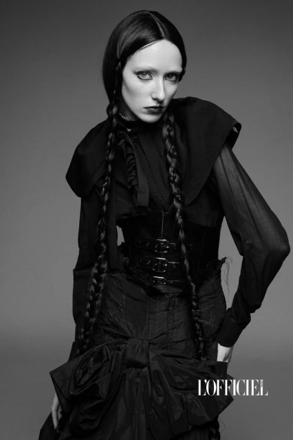 Fashion editorial photo of a slender woman in a black gothic-style outfit and long braids against a gray background, as featured in L'Officiel.