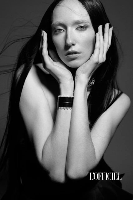 Black and white studio portrait of a woman with long black hair. She poses with her hands framing her face and wearing a wide cuff bracelet.