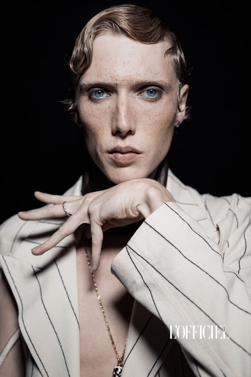 Portrait of a fair-skinned, blue-eyed person with retro blonde wavy hair, wearing a pinstripe jacket against a dark backdrop.