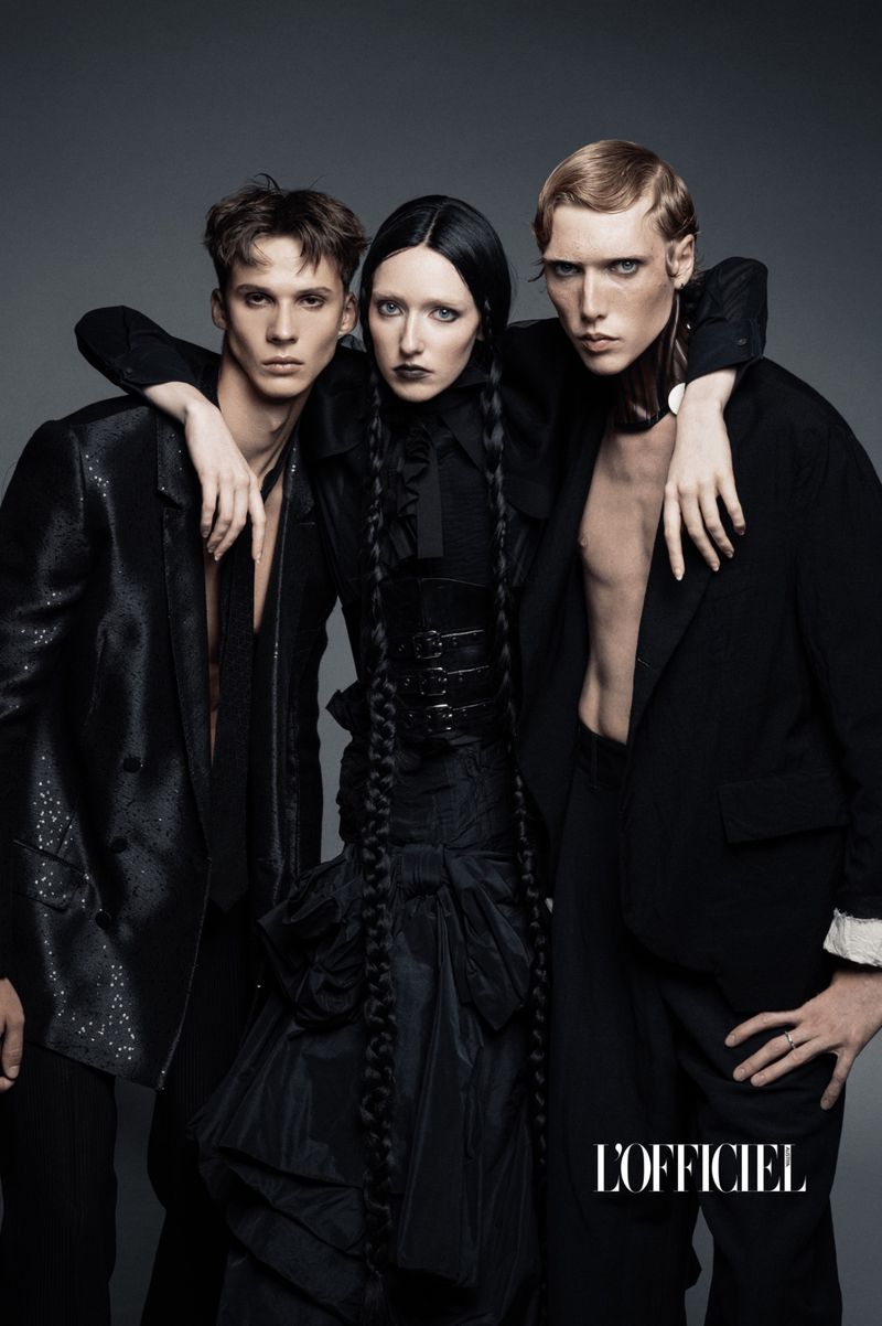 Three fashion models pose in black clothing in a studio setting. The image is a portrait orientation fashion shot.