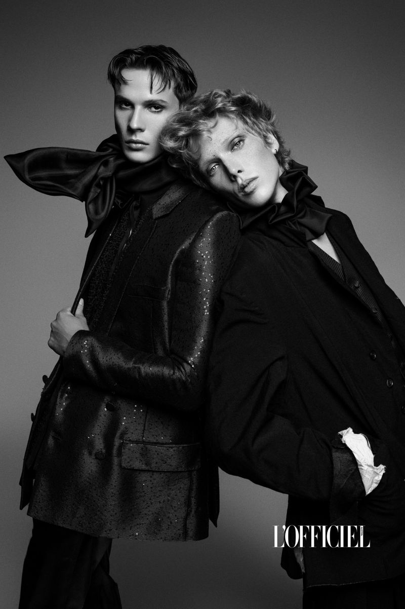 Black and white portrait of two young adult men posing in dark blazers and bow ties against a neutral grey backdrop in a studio.