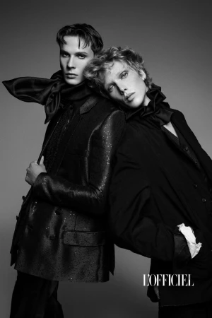 Black and white portrait of two young adult men posing in dark blazers and bow ties against a neutral grey backdrop in a studio.