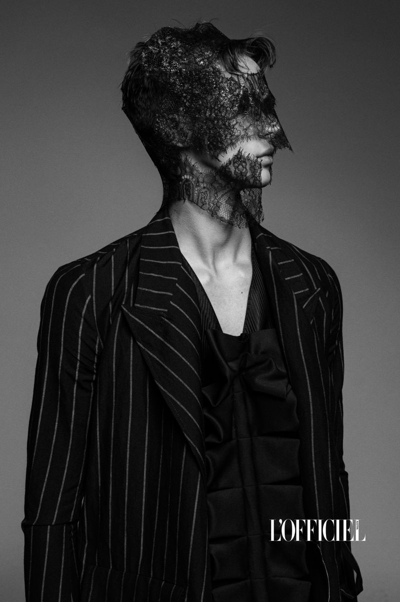 Monochrome fashion shot of a man with face lace, wearing a pinstripe suit and silky top, posing in a photo studio setting.