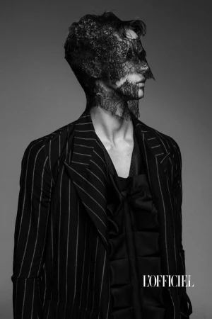 Monochrome fashion shot of a man with face lace, wearing a pinstripe suit and silky top, posing in a photo studio setting.