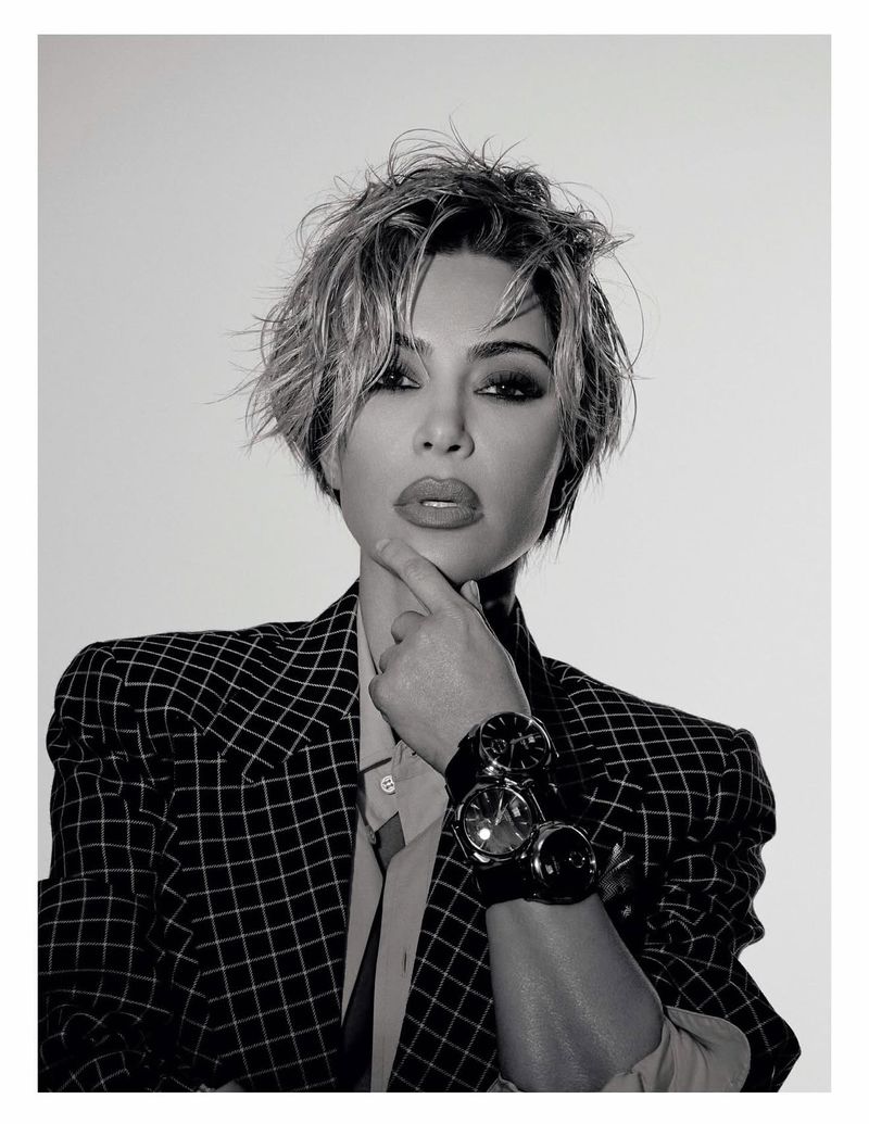 Black and white portrait of an adult female with short hair wearing a checked blazer and multiple watches, gazing directly at the viewer.