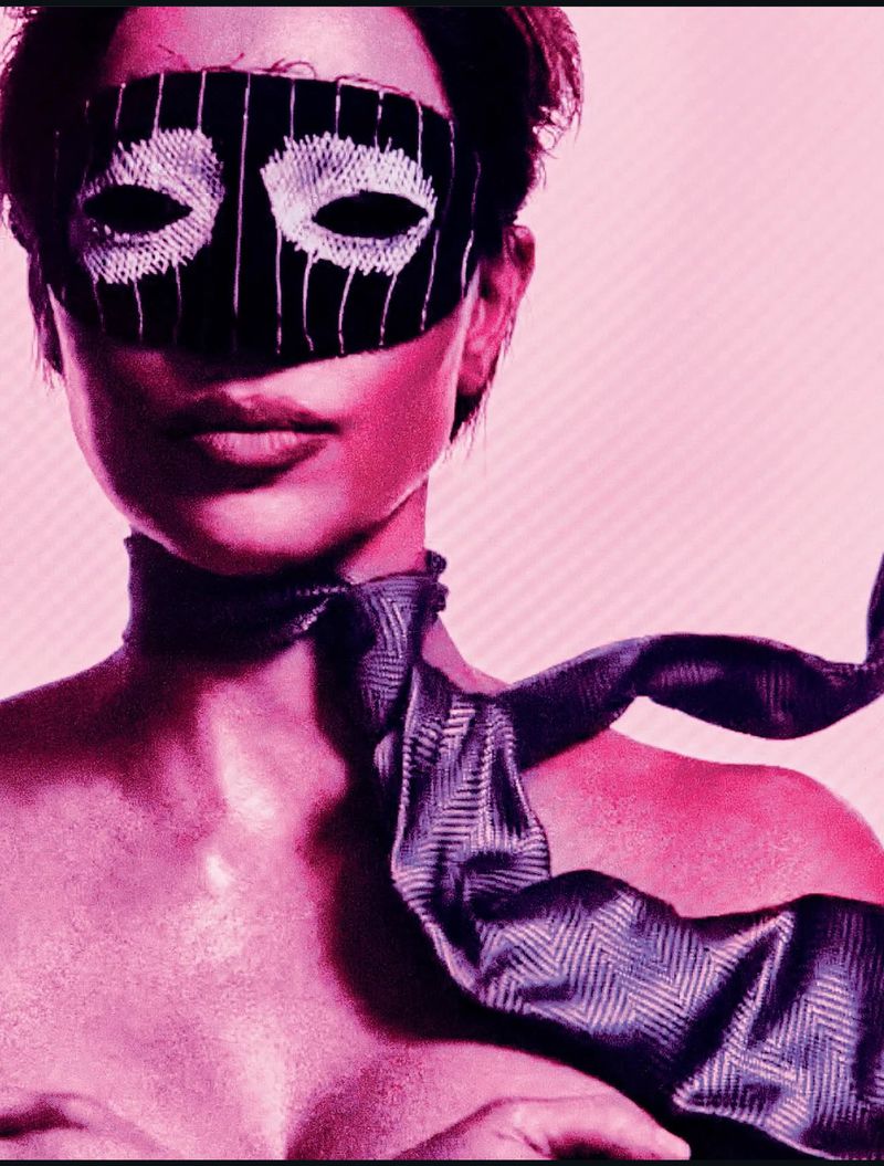 Close-up of a woman wearing a black and white mask and a grey patterned necktie, set against a pink background in a portrait format.