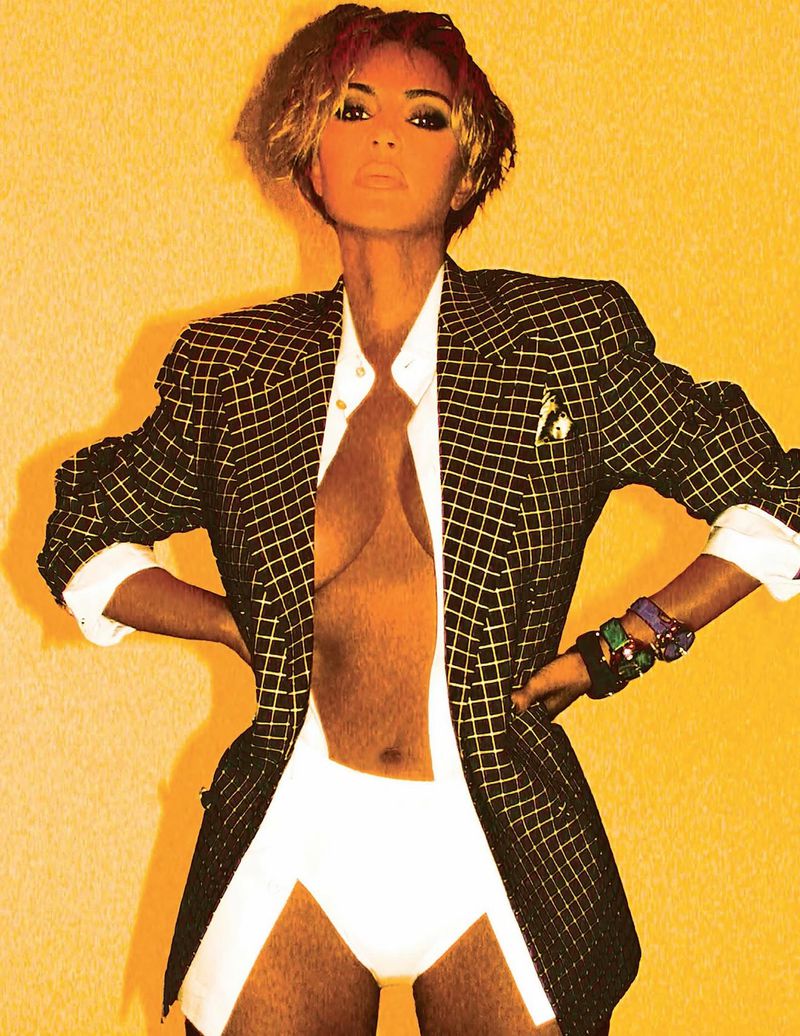 A woman poses in a blazer and bottoms against a yellow backdrop in a fashion shot, showcasing stylish attire and confident posing.