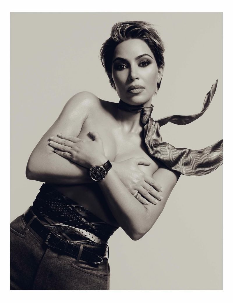 Striking black and white portrait of a topless woman with short hair wearing a scarf, jeans, and a watch against a plain background.