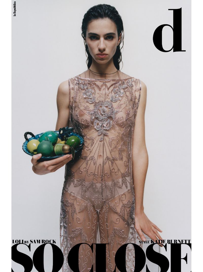 Fashion editorial shot of a woman wearing a sheer beaded dress holding a bowl of fruit against a studio backdrop.
