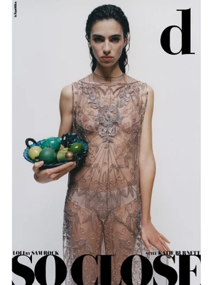Model with wet dark hair wearing a sheer embroidered dress and holding a bowl of green fruit against a plain grey background