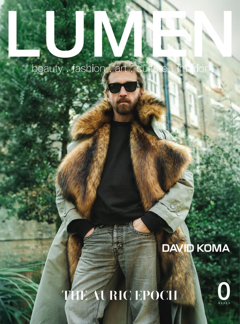 Man wearing sunglasses and a faux-fur trimmed coat standing outdoors against a residential building backdrop
