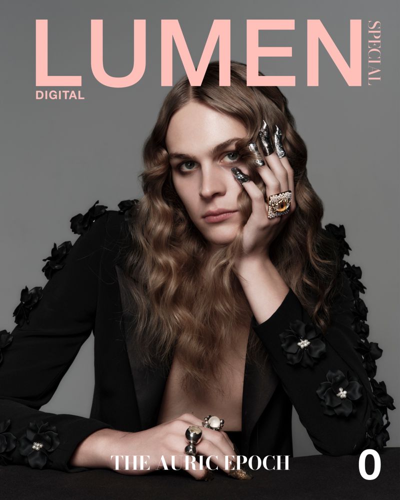 Model with long wavy brown hair wearing a black floral-appliqued jacket and statement rings against a grey seamless background