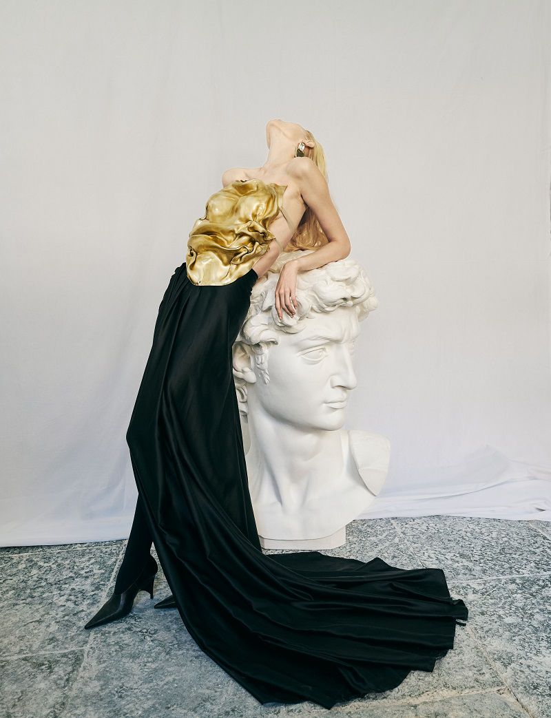 Woman in golden top and black skirt leans on David sculpture in fashion editorial with neutral backdrop, artistic composition, studio lighting.