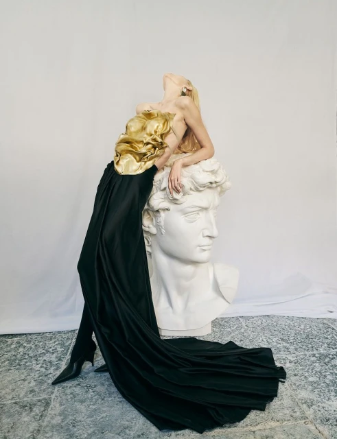 Woman in golden top and black skirt leans on David sculpture in fashion editorial with neutral backdrop, artistic composition, studio lighting.