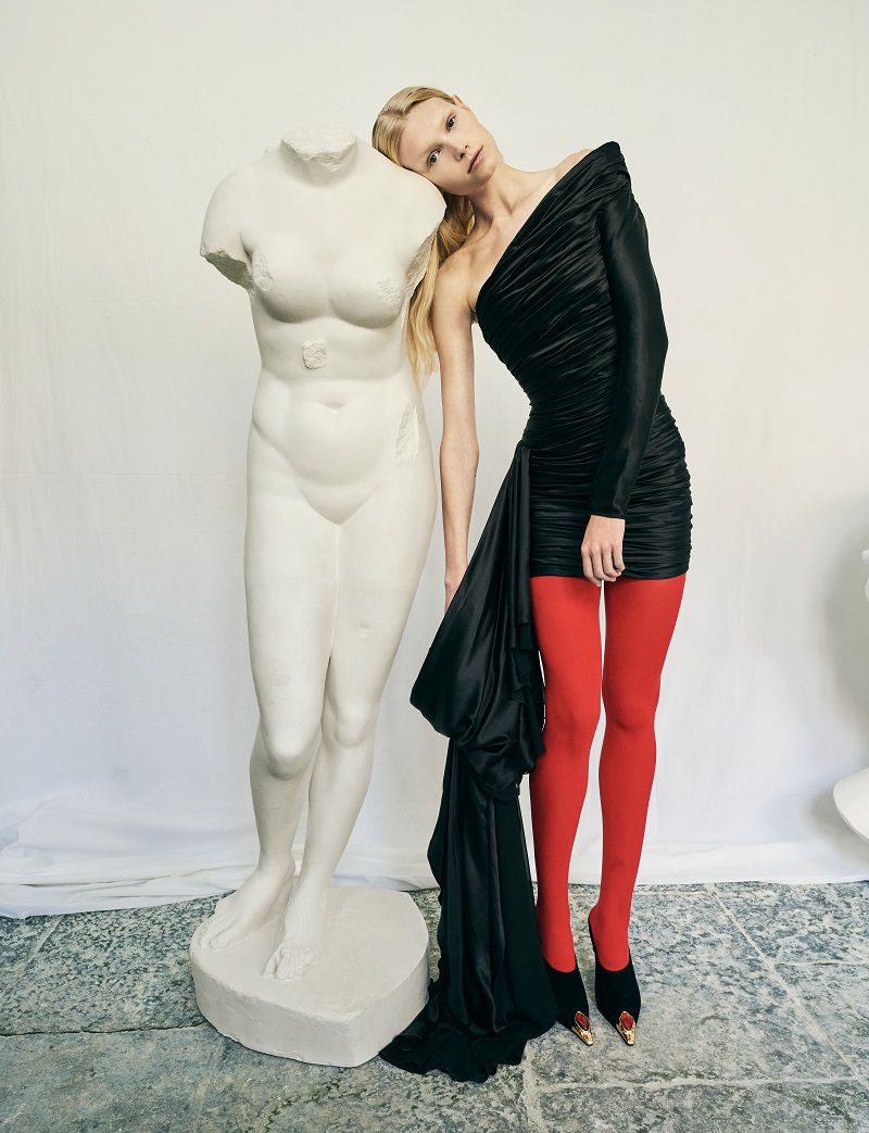 A fair-skinned woman in a black dress and red tights poses next to a white, armless female torso sculpture in a bright indoor setting.