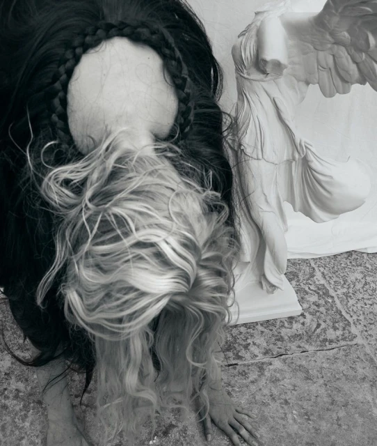 Monochrome portrait of a woman with braided black and blonde hair next to a marble sculpture of Winged Victory of Samothrace.