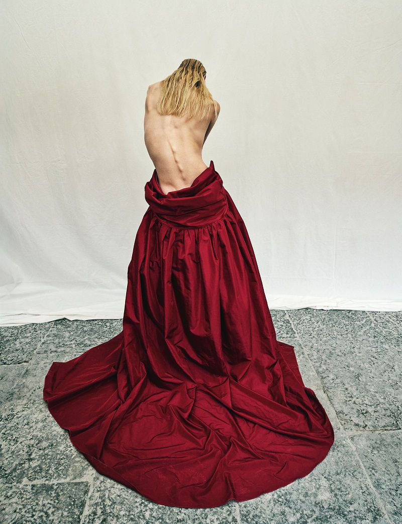 Model with long blonde hair seen from behind, wearing a voluminous red gown draped low on the hips against a white backdrop.