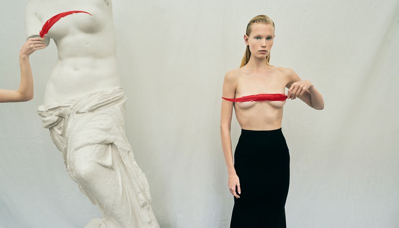 Adult woman in black skirt holds red feather, posing next to white sculpture of woman's torso in a minimalist white studio.