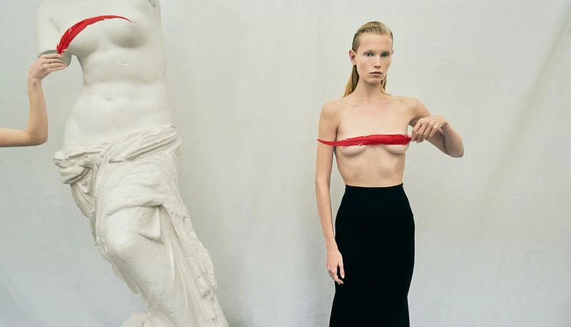 Adult woman in black skirt holds red feather, posing next to white sculpture of woman's torso in a minimalist white studio.