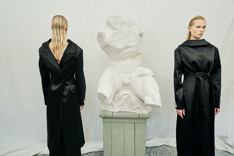 Two fashion models pose in black coats beside a classical bust in a minimalist studio, blending contemporary style with classic art.