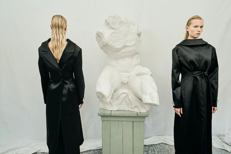 Two fashion models pose in black coats beside a classical bust in a minimalist studio, blending contemporary style with classic art.