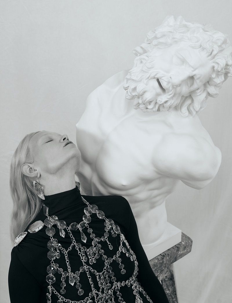 Woman in black turtleneck and crystal jewelry poses next to a marble bust against a white background in a studio fashion shoot.