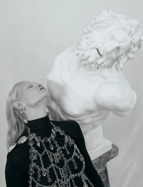 Woman in black turtleneck and crystal jewelry poses next to a marble bust against a white background in a studio fashion shoot.