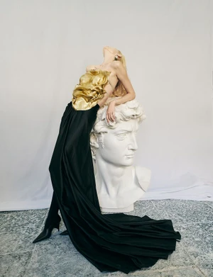 Model Sasha Dom in a sculptural gold top and flowing black draped skirt leaning back against a large classical white David bust sculpture.